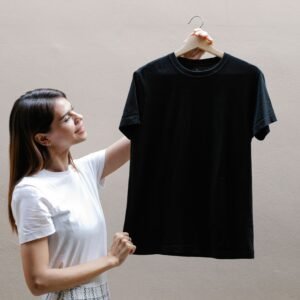 Side view of self assured young woman with long dark hair in stylish outfit smiling while holding hanger with black t shirt against gray wall